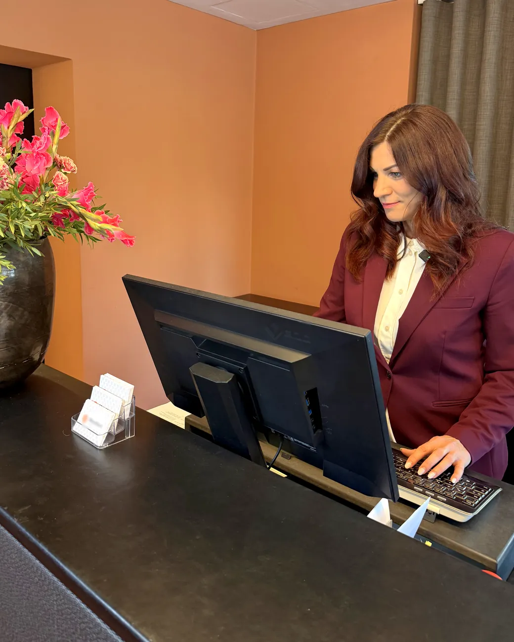 Nina Fleisch, Gründerin der TALAVY Social Suite, lächelt an einer Hotelrezeption und tippt am Computer, daneben ein Strauß pinker Blumen
